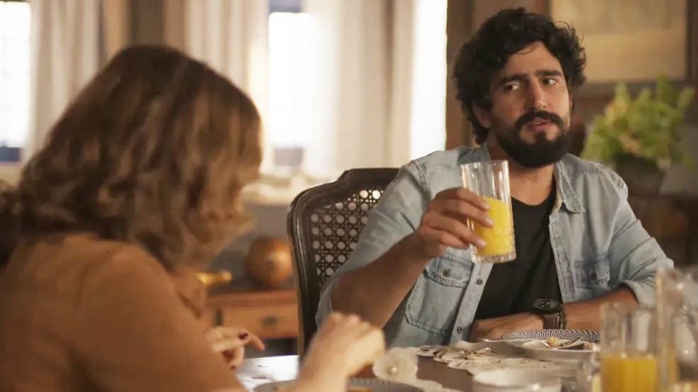 Cena de Mar do Sertão em que Tertulinho aparece segurando um copo com suco de laranja e olhando para Deodora na mesa de jantar